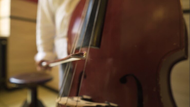 extreme close up of weary used scratched cello bass being played slowly by a professional musician inside building with wooden walls little round seat next to him man wearing white shirt grey pants