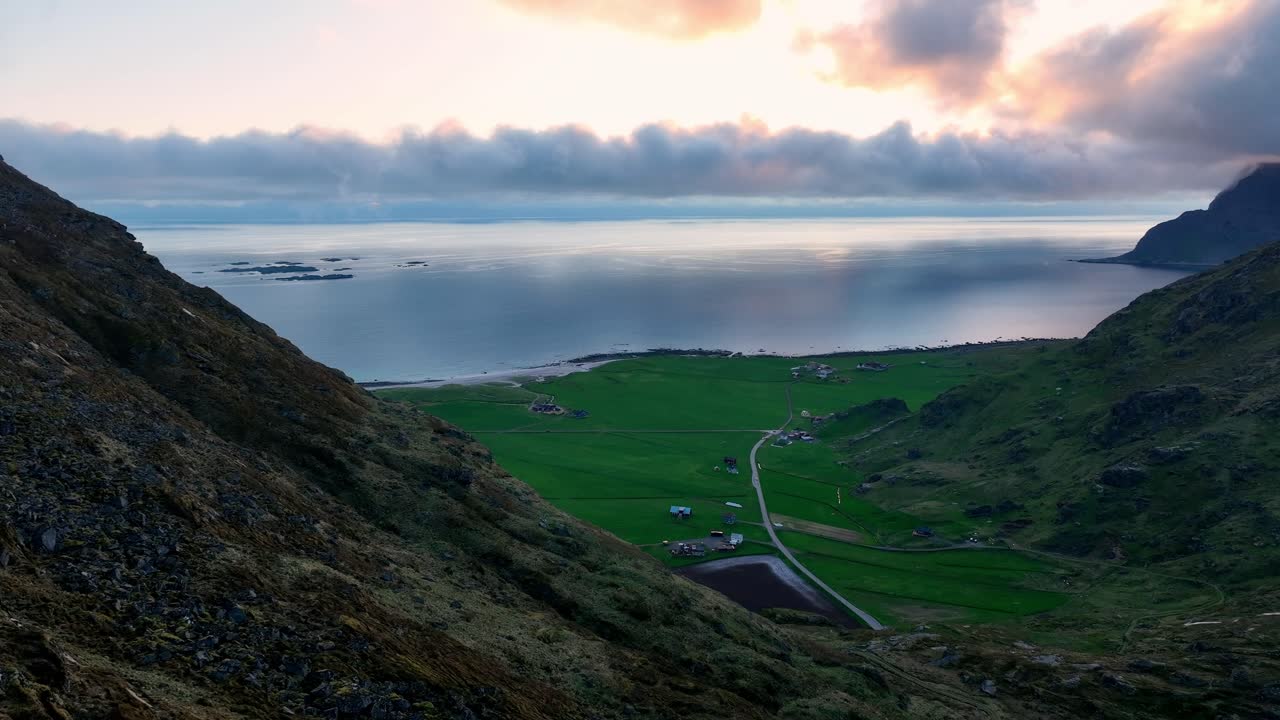 Calm aerial along Mannen cliff with view of Utakleiv beach, green fields and midnight sun