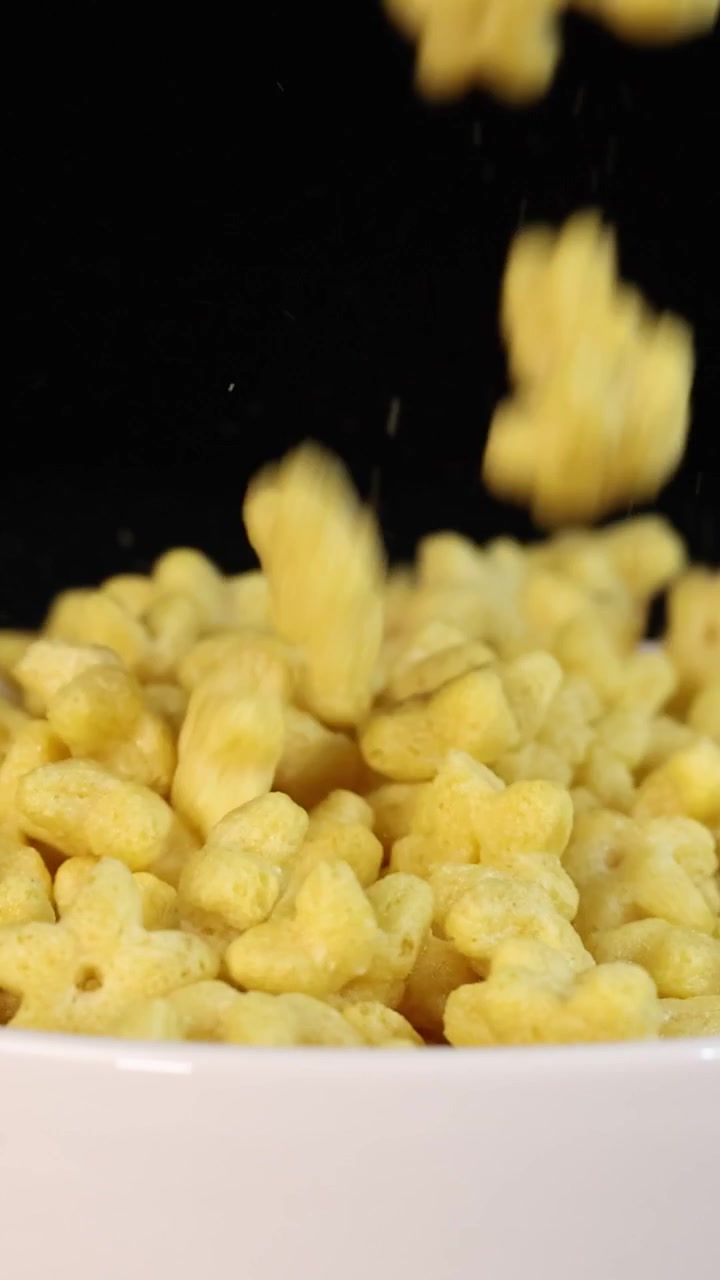 Pezzi di cereali gialli a forma di stella cadono in una ciotola di ceramica bianca su uno sfondo nero.