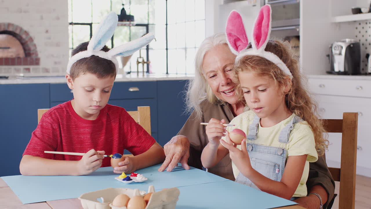 Grandmother With Grandchildren Wearing Rabbit Ears Decorating Easter Eggs At Home Together