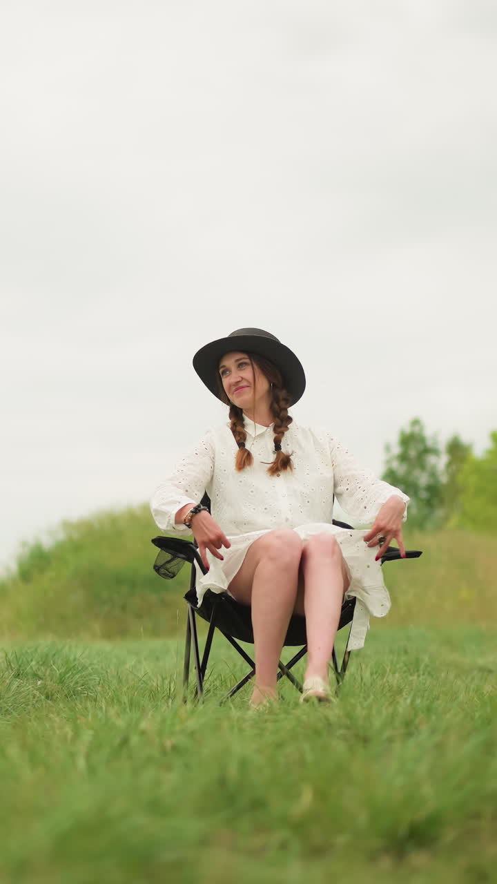 색 드레스와 검은색 모자를 입은 아름다운 여성이 잔디에 있는 의자에 앉아 자신감 있게 포즈를 취하고 있다. 색 셔츠를 입은 남자가 을 들고 그녀를 면밀히 지켜보고 있다.