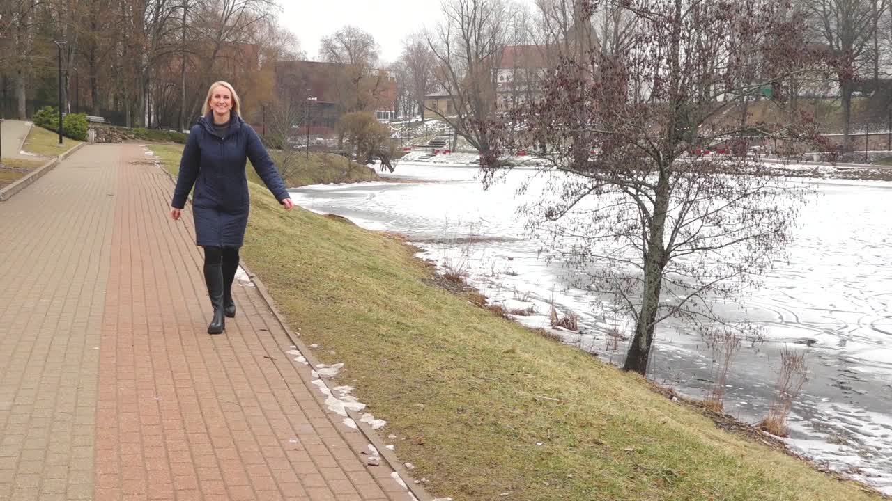 A woman with long blonde hair smiles and speaks while standing outdoors in front of evergreen trees, dressed warmly in a navy coat and beige scarf.