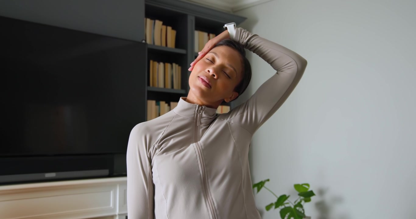 Mujer afroamericana haciendo estiramientos de cuello y hombros en casa, calentando con chaqueta y reloj inteligente.