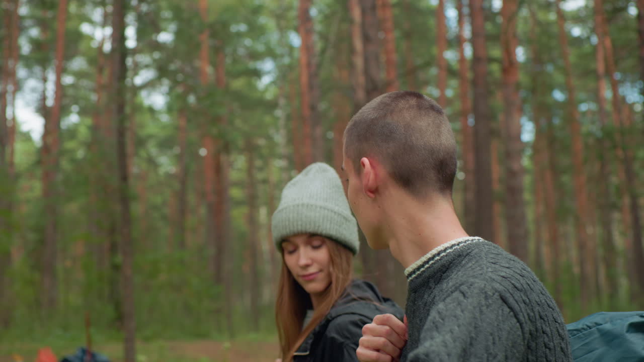 Side view of friends walking through forest as boy looks around while holding bag strap and girl glances ahead, surrounded by tall trees and serene greenery