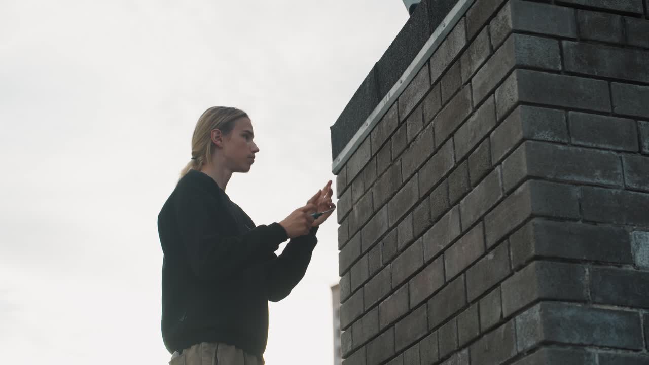 Vaalea mies lukee tekstiä nojaten seinään. Kaupunkilainen hupparissa tutkii puhelinta tiiliseinän vieressä ulkona. Vaaleaihoinen mies vaaleilla hiuksilla käyttää laitettaan ulkona urbaanin tiiliseinän vieressä