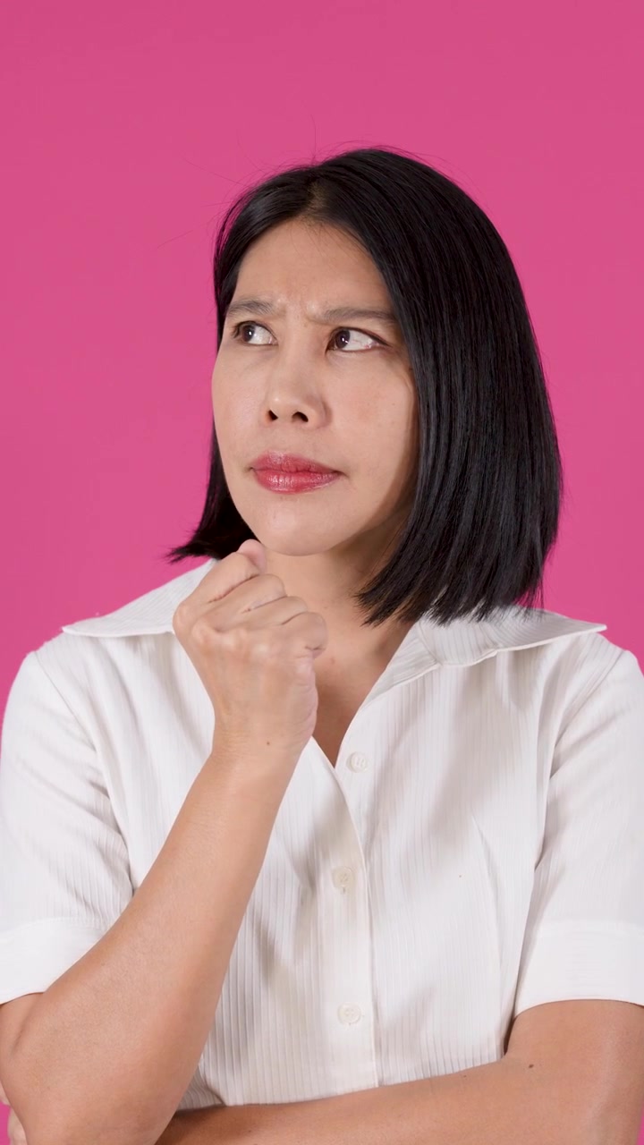 Medium shot of a frustrated Asian woman contemplating with hand on chin against pink background