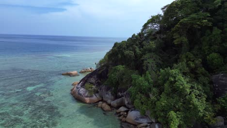 Turquoise-ocean-water-meeting-a-rocky-shore-with-a-lush-green-jungle-forest-on-a-tropical-island