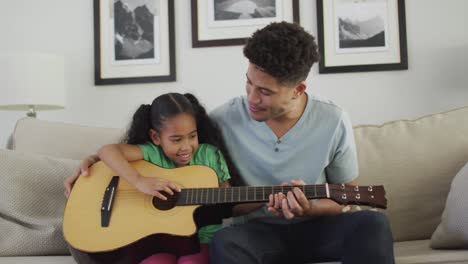 happy biracial father and daughter sitting on sofa playing guitar