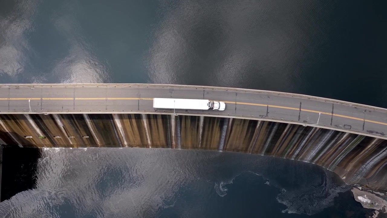 Water flowing through the spillway of curved dam structure and hydroelectric Generating Station infrastructure surrounded by deep blue river water, birds eye aerial