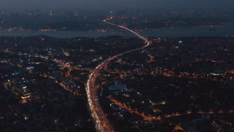 Highway-Freeway-Road-going-through-Istanbul-City-with-Red-Illuminated-Bosphorus-Bridge-in-the-distance-at-Night,-Aerial-Wide-View