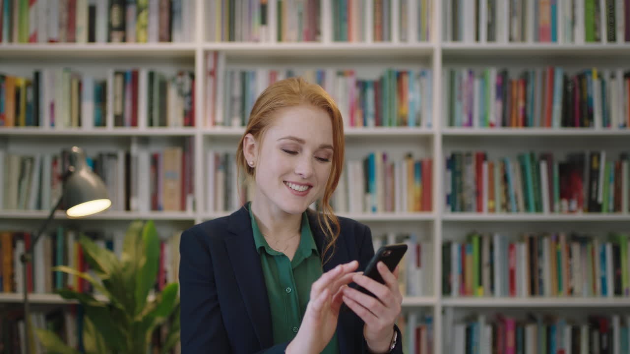 portrait of beautiful young red head business woman executive smiling enjoying texting browsing using smartphone mobile app in library background