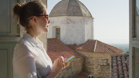 Hermosa-Mujer-Usando-Un-Teléfono-Inteligente-Navegando-En-Línea-Enviando-Mensajes-De-Texto-Disfrutando-Compartiendo-Experiencias-De-Vacaciones-De-Verano-Relajándose-En-El-Balcón-De-Una-Villa-Rústica-Con-Gafas-De-Sol