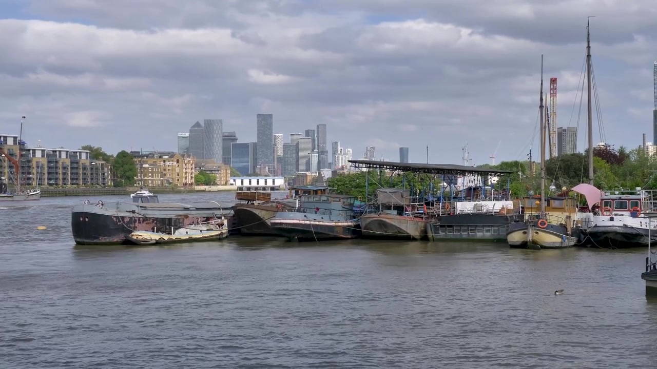 Case galleggianti e chiatte usurate ormeggiate a più livelli sul fiume con un ponte giardino coperto, contro uno skyline di torri bancarie in vetro sotto nuvole grigie.