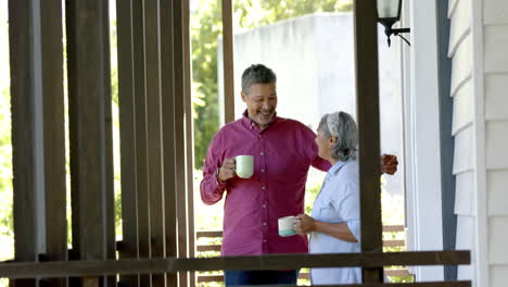 Feliz-Pareja-Birracial-De-Ancianos-Abrazándose-En-La-Ventana,-Hablando-Y-Bebiendo-Té