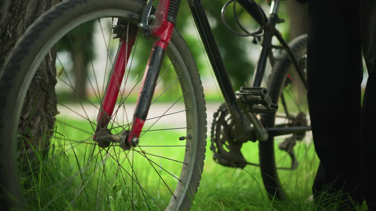 주차된 자전거의 페달에 발을 고 잔디에 있는 나무에 기대어 자전거와 부품에 초점을 맞추고 위치를 확인하는 사람의 클로즈업, 흐릿한 초록색 배경