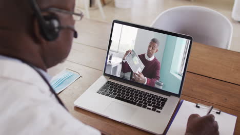 african american male doctor wearing phone headset taking notes while having a video call on laptop