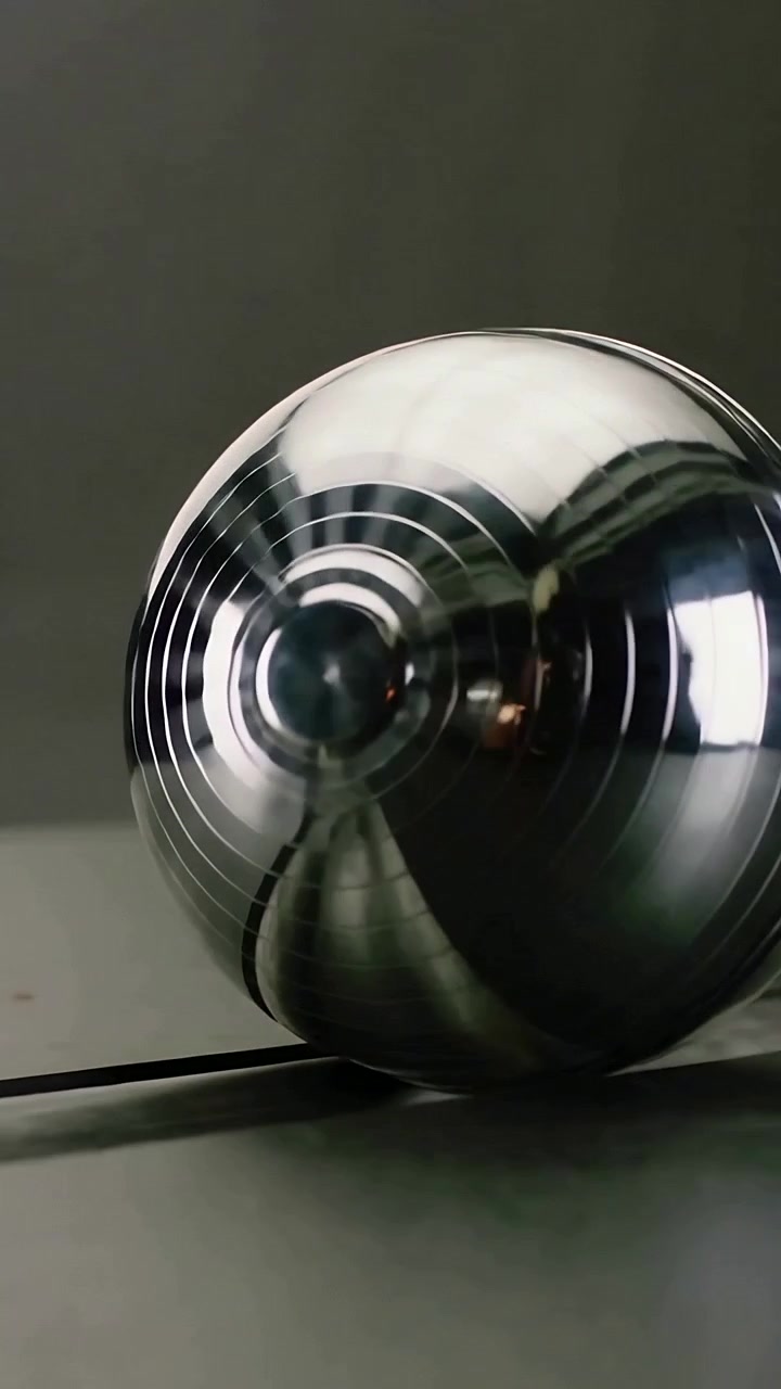 Vídeo vertical: Bola cromada ranhurada girando e rolando pela emenda da mesa, mostrando reflexos de luz.