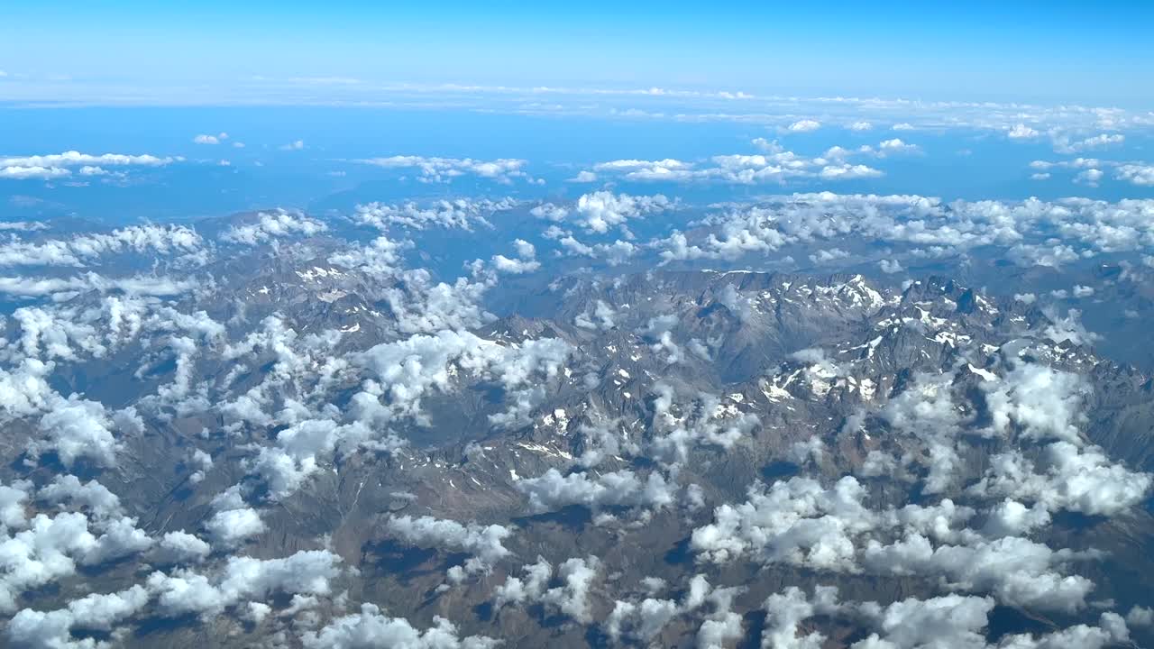 An elevated aerial left side view of the Alps mountains with almost no snow and with scattered low clouds, taken from an airplane cockpit in a summer morning