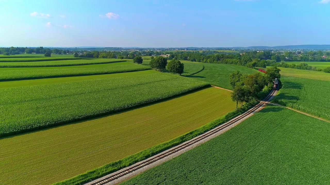 Scenic Aerial View of Lush Green Farm Fields With a Steam Train, Blowing Smoke and Steam Approaching Around a Curve on a Sunny Day