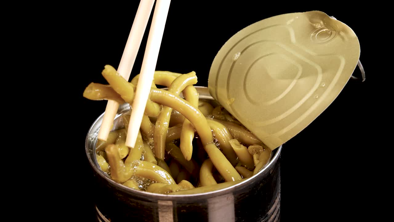 Chopsticks grasp and lift haricot beans from an open canned food container against a black background, with even studio lighting and close-up framing