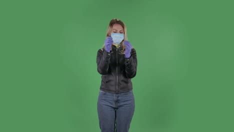 의료 마스크를 입은 아름다운 젊은 여성의 초상화는 카메라를 보고 고무 장갑을 착용합니다. 검은색 재과 청바지를 입은 느슨한 머리카락을 가진 금발은 스튜디오에서 녹색 화면에 있습니다. 건강 보호 코로나 바이러스 개념