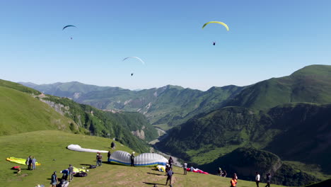 Paragliders-take-off-from-the-side-of-a-mountain,-while-others-are-setting-up-their-equipment