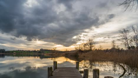 el impresionante resplandor del amanecer se extiende a través de las nubes que se reflejan en el agua del lago al final del muelle de madera con cañas y casa de granja, pfäffikersee en suiza