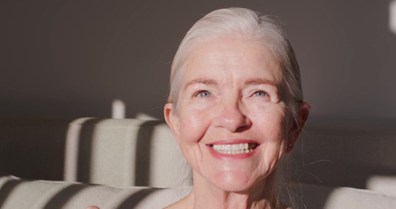 Senior woman squinting from bright sunlight, turning head forward and smiling on sofa in tank top
