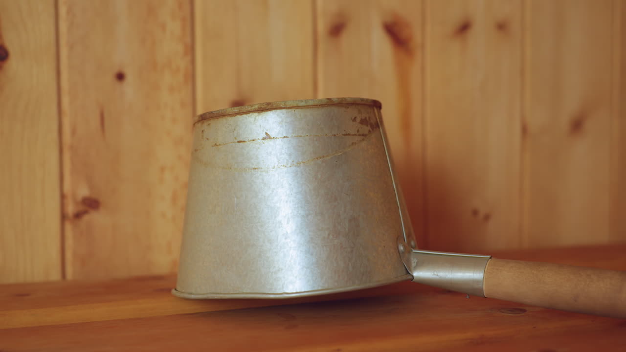 Close up of worn metallic ladle with long wooden handle resting on smooth wooden sauna bench, surrounded by natural vertical wood paneling in warm lighting