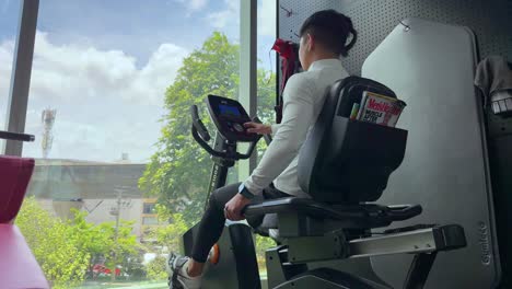 a man works out on a recumbent bike in a gym with large windows, under bright natural lighting