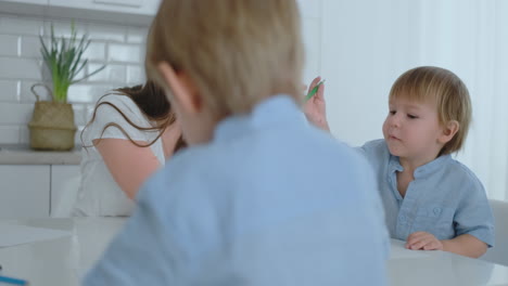 two children of boys draw with his mother sitting in the kitchen.