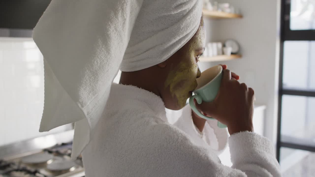 Felice donna afroamericana con una maschera di bellezza sul viso che beve caffè in cucina.