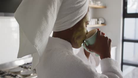 happy african american woman with beauty mask on face, drinking coffee in kitchen