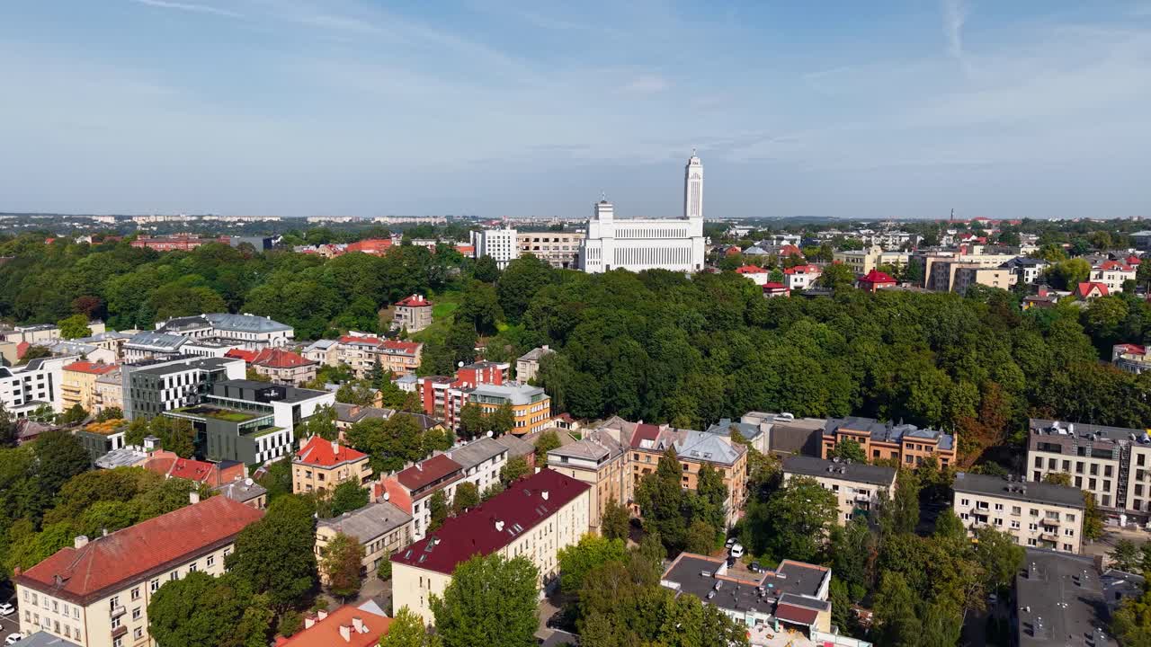 Aerial view of Kaunas city in Lithuania with residential and commercial buildings, lush greenery, and the prominent Church of the Resurrection in the distance under a clear blue sky