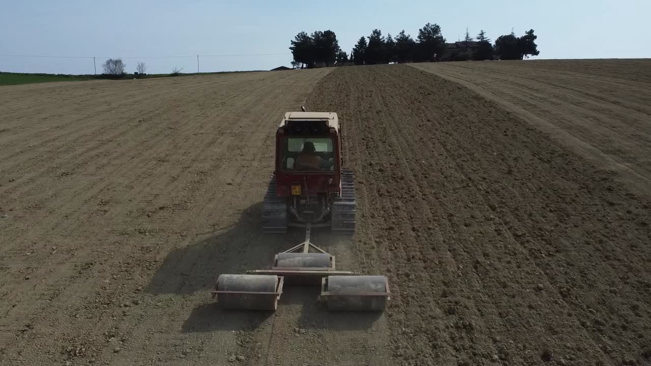 Drone follow red tractor rolling the soil creating better seedbed for germinating seeds, aerial agricultural farming natural soil land