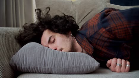 A-tired-brunette-guy-with-curly-hair-in-a-checkered-shirt-falls-on-a-gray-pillow-at-home-after-a-hard-day-at-work