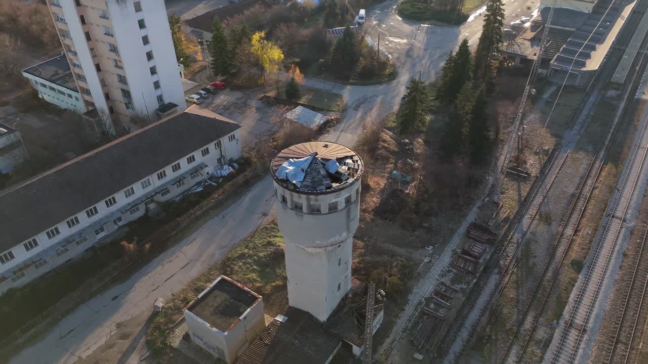 Aerial drone view of an old watchtower located beside railway tracks in Germany. Surrounded by buildings and trees, this scene captures an abandoned structure in a quiet, rural area at sunset.
