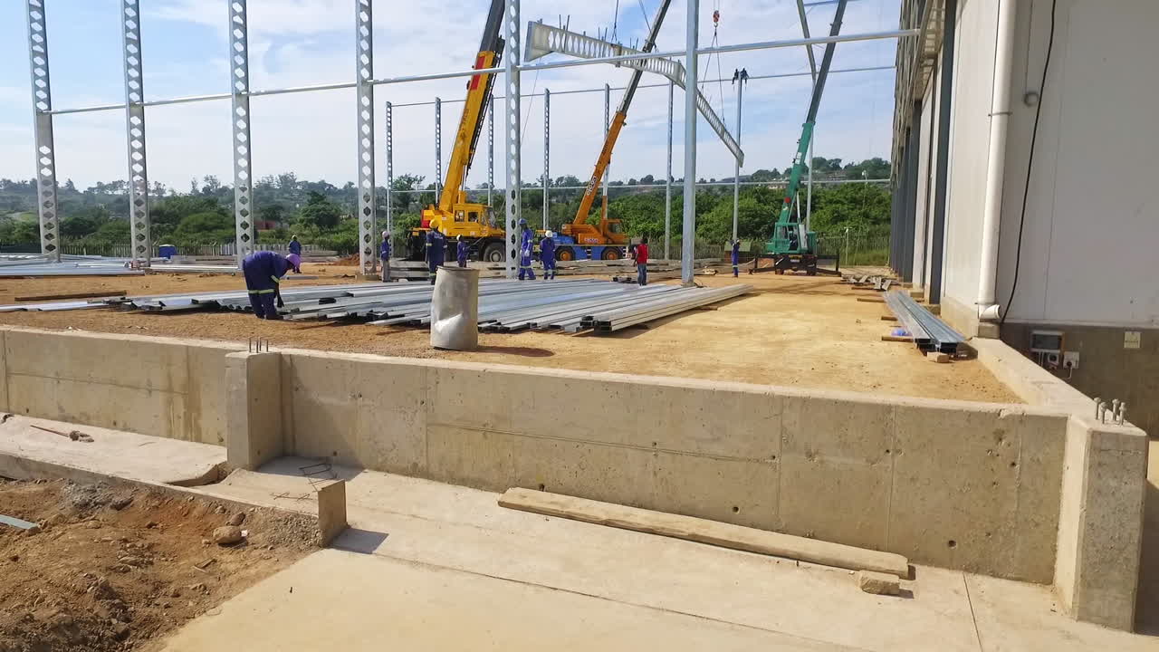 drone, los trabajadores llevan una pesada viga de acero mientras las grúas se levantan en el sitio de construcción