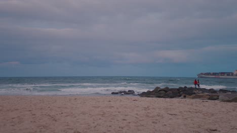 A-large-cargo-ship-sails-by-on-the-horizon-of-a-nearby-sandy,-white-beach-whilst-the-sky-brews-a-dangerous-storm-above,-giving-dark,-moody-tones-to-the-clouds-and-ocean-water