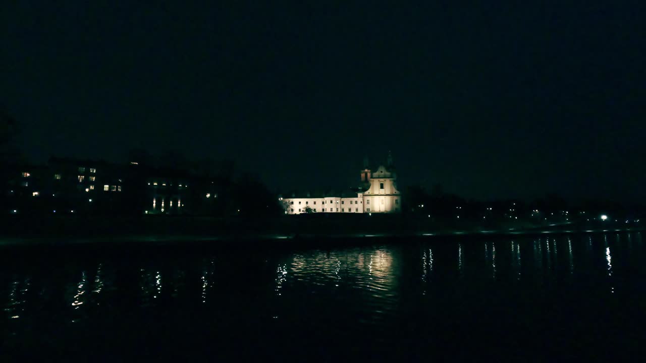 Le luci della città brillano di notte riflettendosi sull'acqua calma. Un edificio storico si erge maestoso lungo la riva del fiume. La vista cattura un momento di bellezza serale.