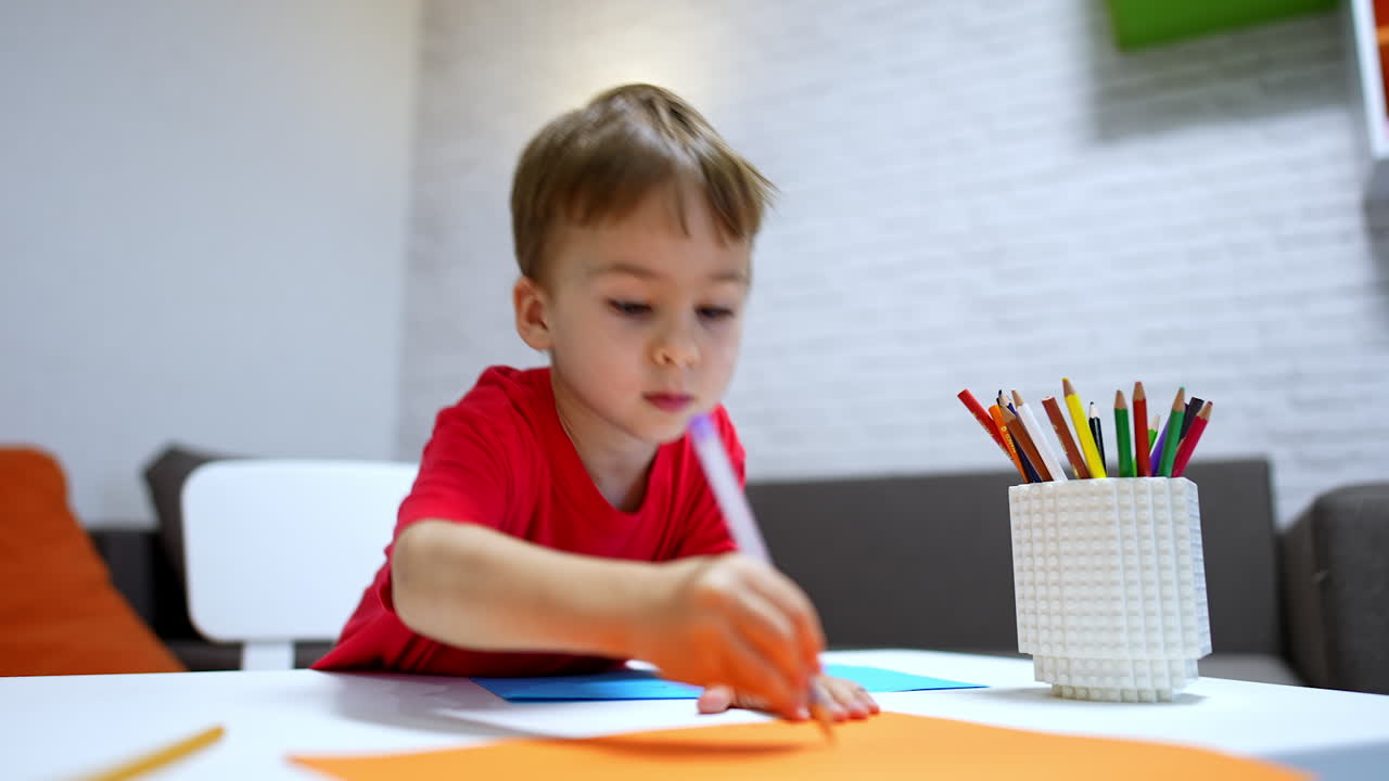 Talkative Caucasian toddler sitting at the desk draws with a pen. Cheerful kid draws on the color paper.