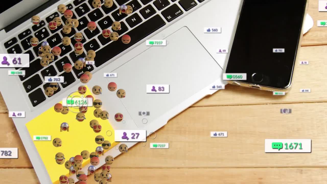 Animace ikon sociálních médií a obličejových emotikonů na pozadí notebooku a chytrého telefonu na dřevěném stole.