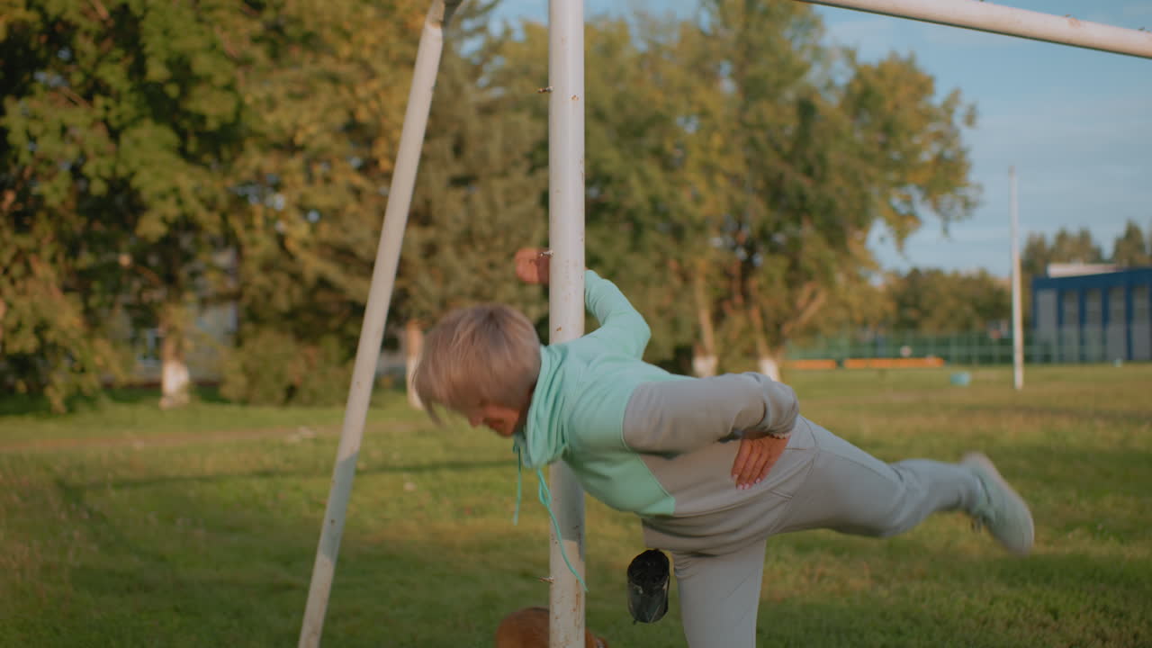 Female trainer holding post performing waist exercise during sunset while puppy tied nearby on grassy field surrounded by trees and building in background demonstrating outdoor fitness