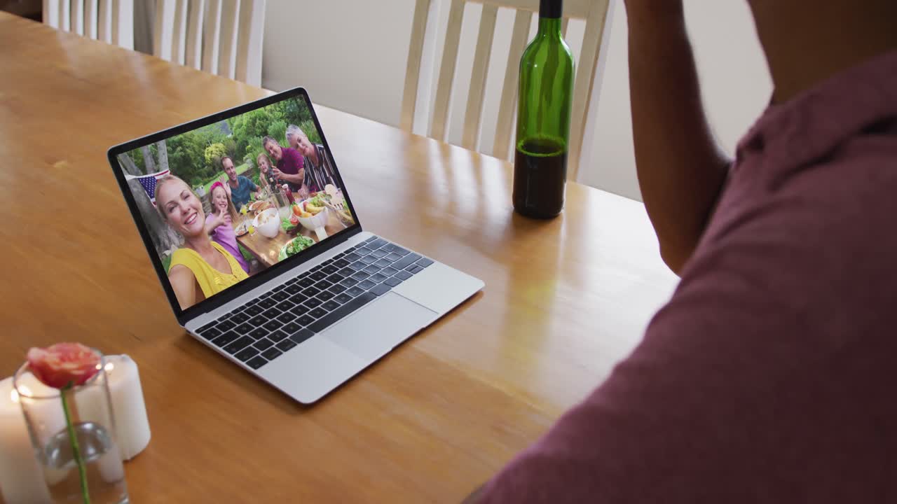Busto di uomo afroamericano che beve vino durante una videochiamata al laptop a casa.