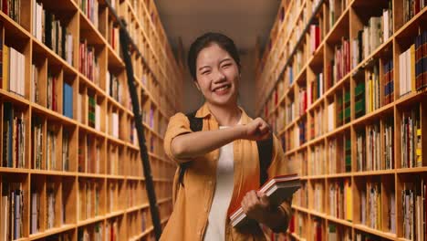 asian woman student with a backpack and some books touching her chest then smiling and pointing to camera being proud of herself in the library