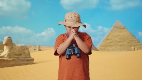 asian boy with a hat and binoculars praying for something while traveling in giza pyramid. boy researcher examines something, travel tourism adventure concept