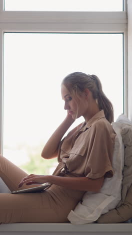 young freelancer woman with ponytail leaned on pillow works on project via modern laptop sitting at windowsill in room at home slow motion