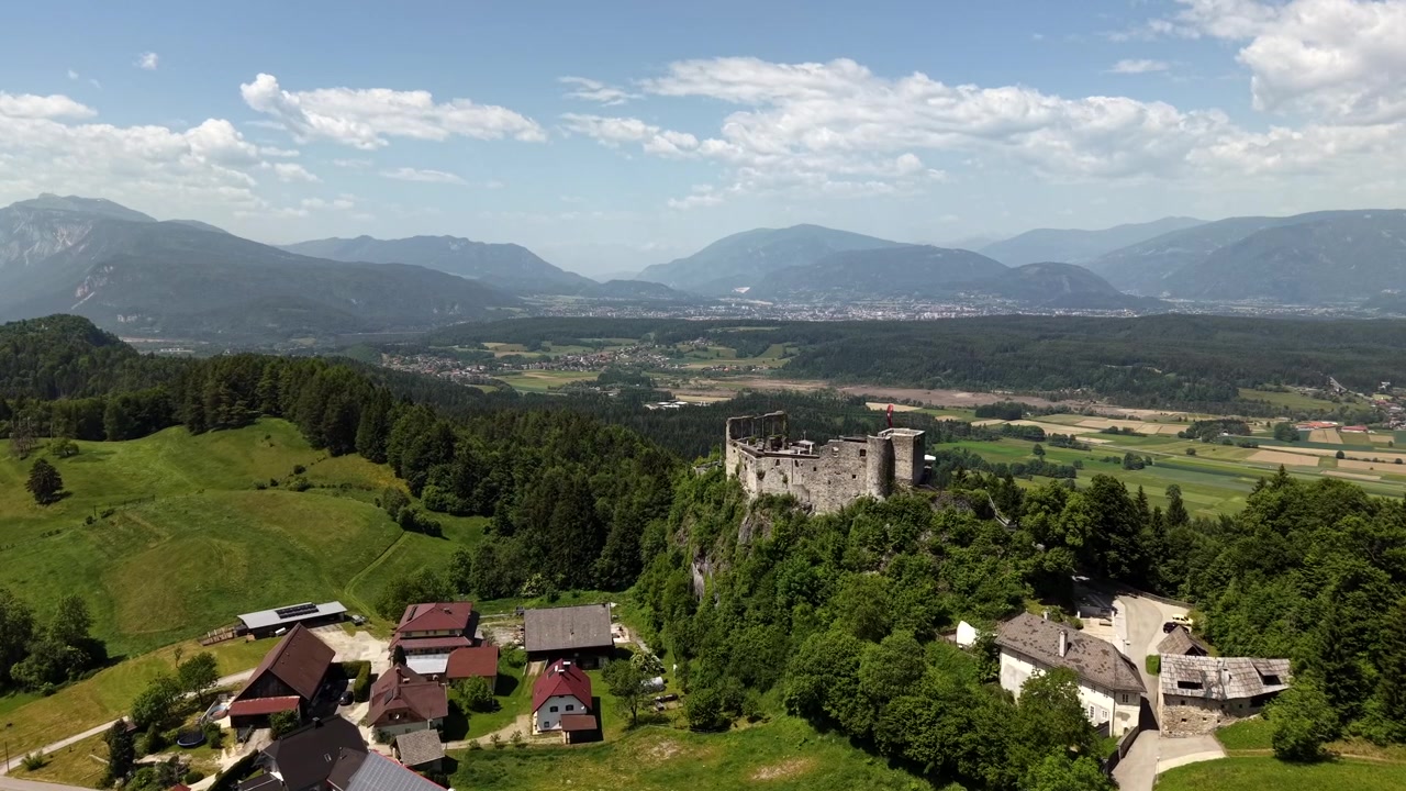 Le rovine del Castello di Finkenstein coronano una collina boscosa sopra un piccolo villaggio austriaco, con una verde valle alpina che si estende verso distanti montagne nebbiose vicino alle acque del Faaker See. Ripresa aerea con drone in avvicinamento.