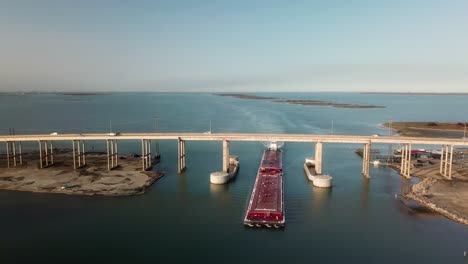 Aerial-view-of-a-large-red-barge-moving-along-the-Gulf-Intercoastal-Waterway-and-under-JFK-Memorial-Causeway-bridge