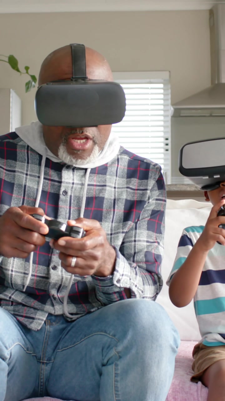 Video verticale: Anziano uomo afroamericano e un ragazzo che giocano a un gioco VR sul divano, tenendo i controller.